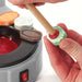 close up of hands holding a dop stick o a green gemstone with melted dop wax and in the background is a dop station with quartz and agates
